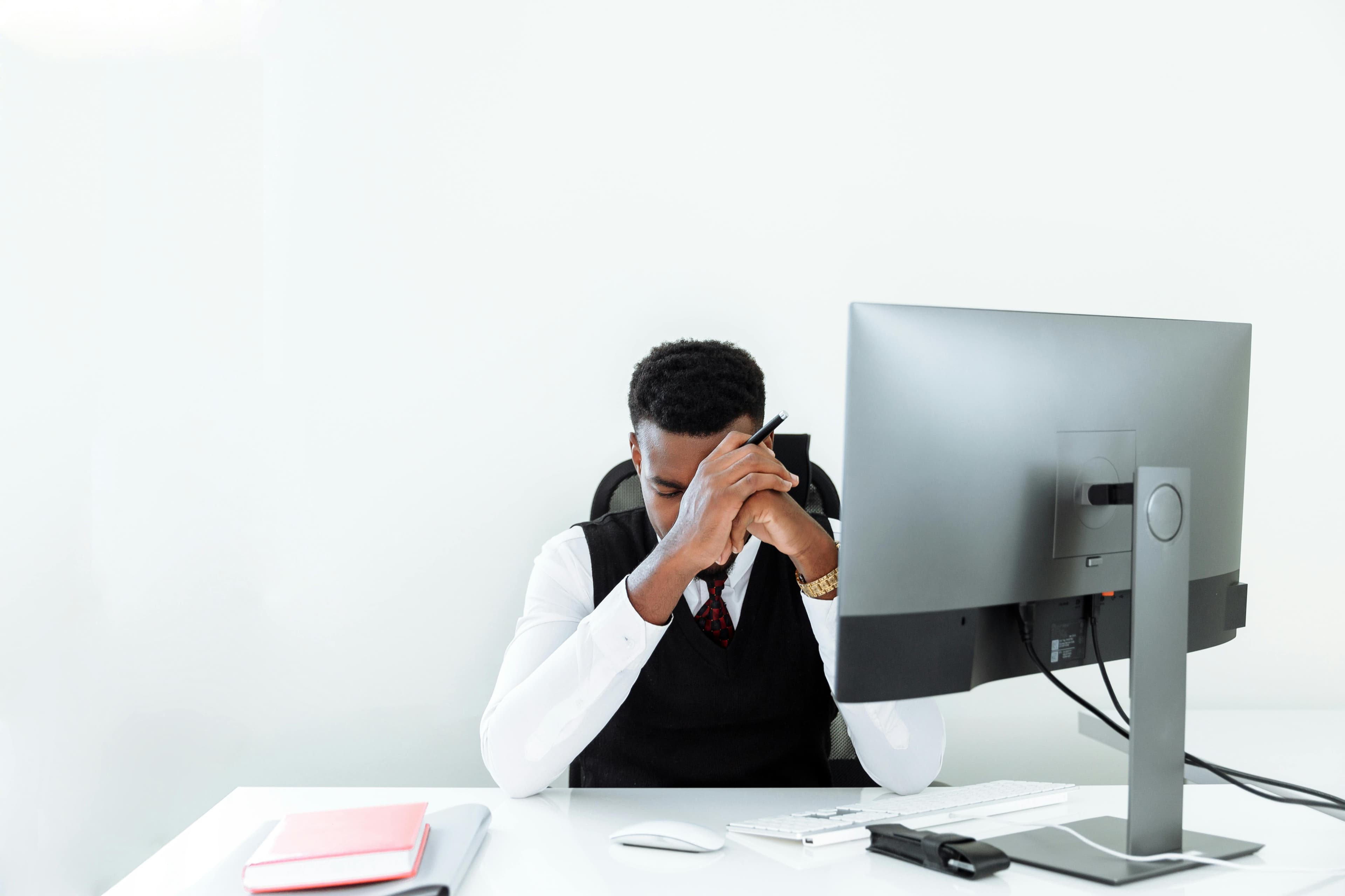 A businessman sitting at a desk, appearing stressed while working at a computer in a bright office